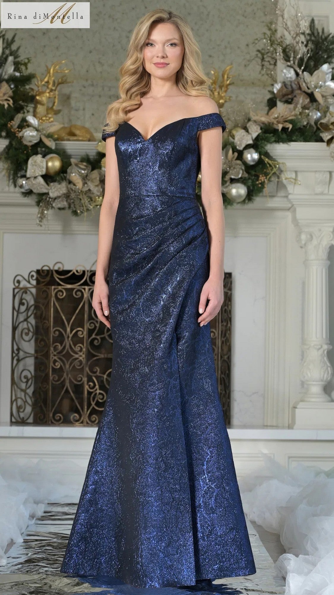 A woman stands indoors wearing the Rina Di Montella RD3364 long formal dress in royal, featuring an off-the-shoulder design and subtle metallic pattern. Festive decorations and a glowing fireplace appear in the background.